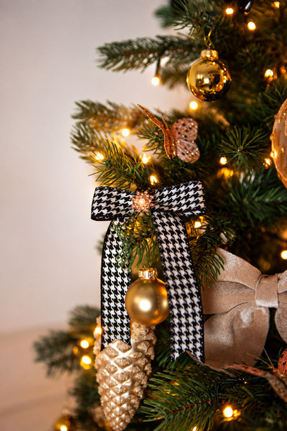 Detail van Christmas Couture kerstboomversiering met een zwart-wit pied-de-poule strik, gouden kerstballen, een gouden dennenappelhanger en warme kerstverlichting.