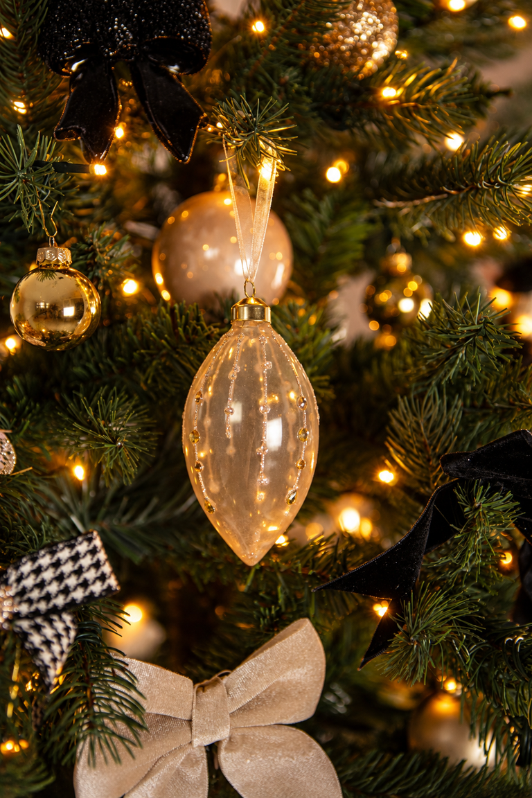 Detail van Christmas Couture kerstboomversiering met een langwerpige glazen kersthanger, gouden en champagnekleurige kerstballen, zwarte en beige strikken en warme kerstverlichting.
