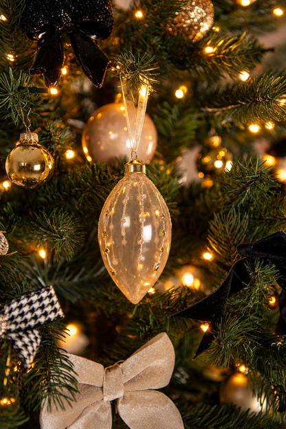 Detail van Christmas Couture kerstboomversiering met een langwerpige glazen kersthanger, gouden en champagnekleurige kerstballen, zwarte en beige strikken en warme kerstverlichting.