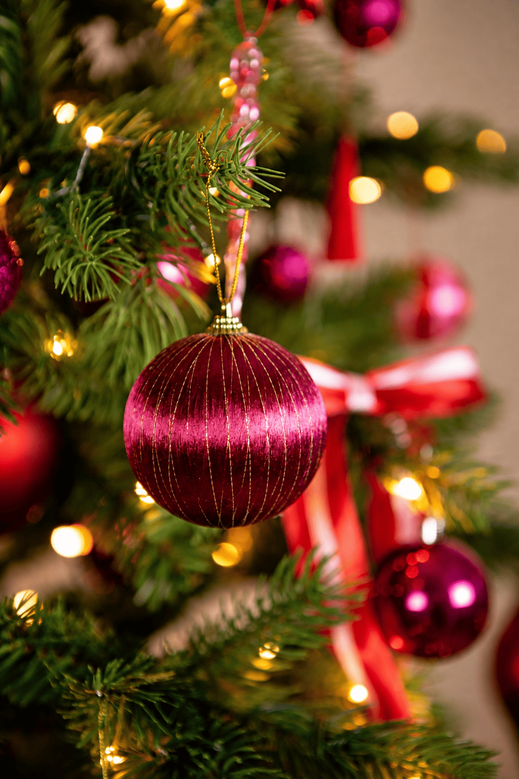 Detail van A Jolly Christmas kerstboomversiering met een roze kerstbal met gouden glitterstrepen, rood-wit gestreepte strikken en warme kerstverlichting.