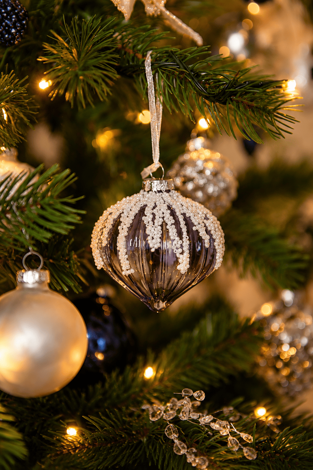 Detail van Snow White Christmas kerstboomversiering met een donkerblauwe glazen kersthanger met witte ijsachtige versiering, zilveren kerstballen en warme kerstverlichting.