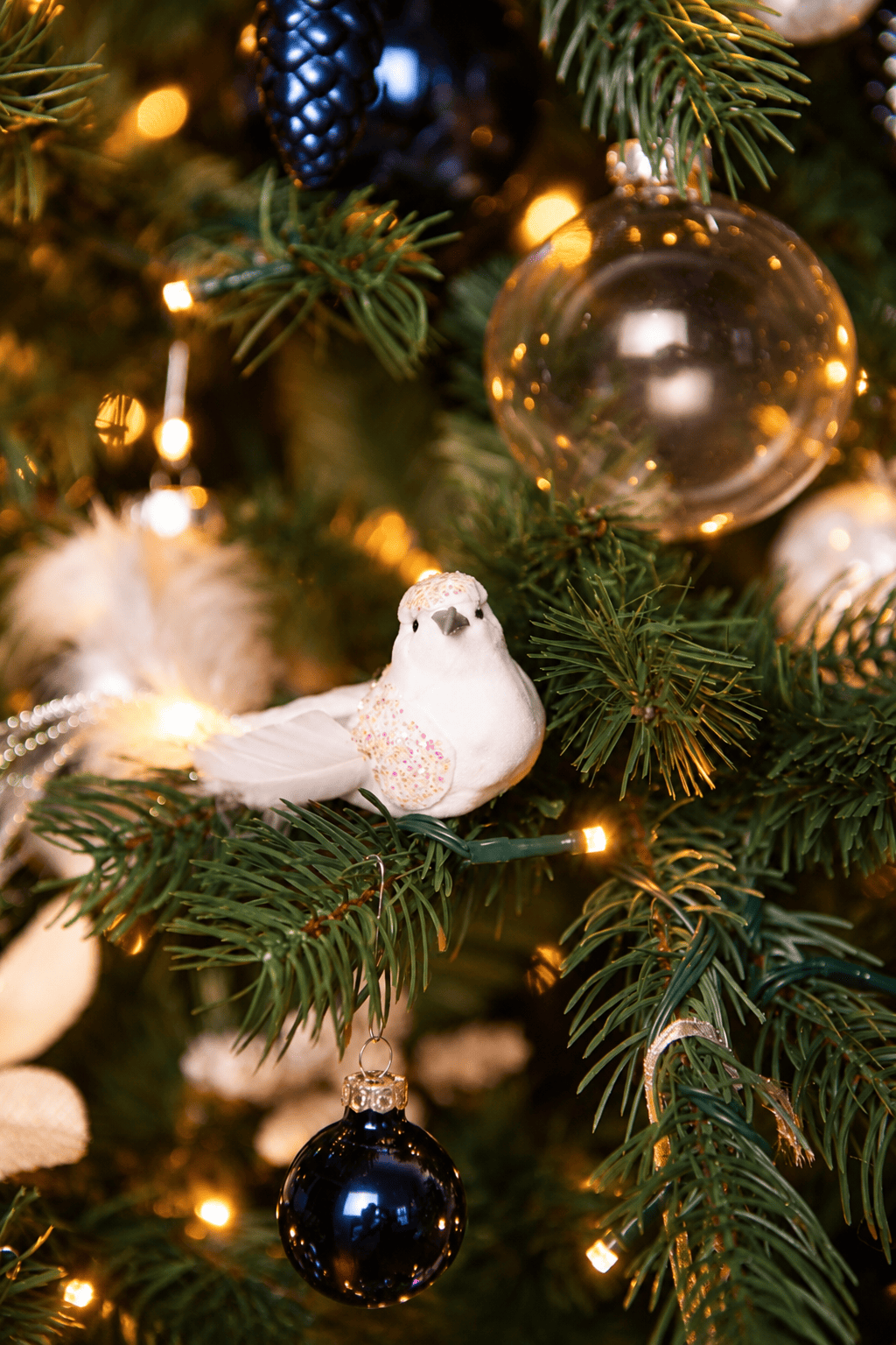 Detail van Snow White Christmas kerstboomversiering met een witte siervogel, transparante en donkerblauwe kerstballen, een blauwe dennenappelhanger en warme kerstverlichting.