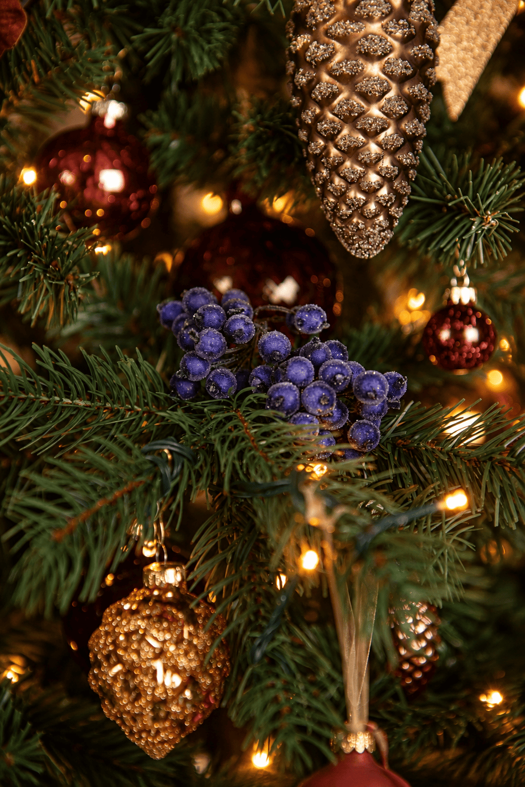 Detail van Warm Christmas Wishes kerstboomversiering met paarse besjes, gouden dennenappels, koperkleurige kerstballen en warme kerstverlichting.