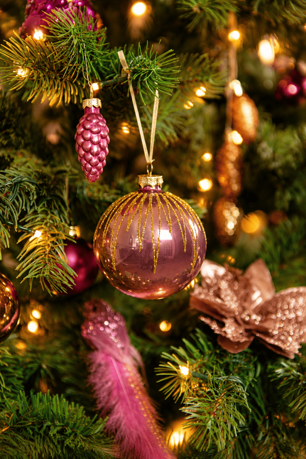 Detail van Warm Christmas Wishes kerstboomversiering met een roze kerstbal met gouden glitters, roze dennenappel, sierveer en warme kerstverlichting.
