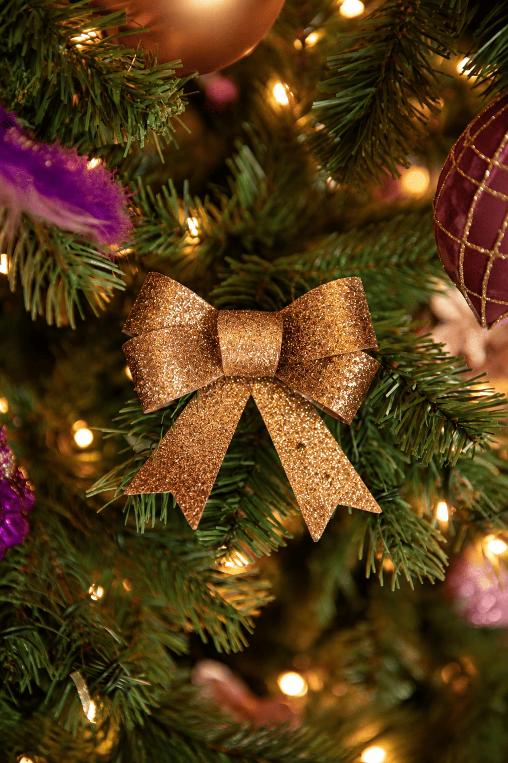 Detail van Warm Christmas Wishes kerstboomversiering met een gouden glitterstrik, paarse sierveer, roze kersthanger en warme kerstverlichting.
