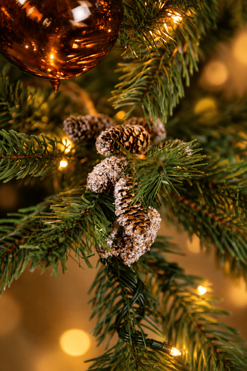 Detail van Warm Christmas Wishes kerstboomversiering met besneeuwde dennenappels, een koperkleurige kerstbal en warme kerstverlichting.