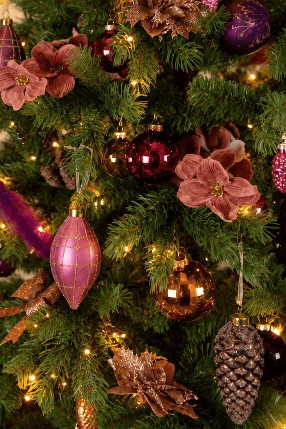 Detail van Warm Christmas Wishes kerstboomversiering met roze en paarse kerstballen, roze kerstbloemen, gouden accenten, een dennenappel en warme kerstverlichting.